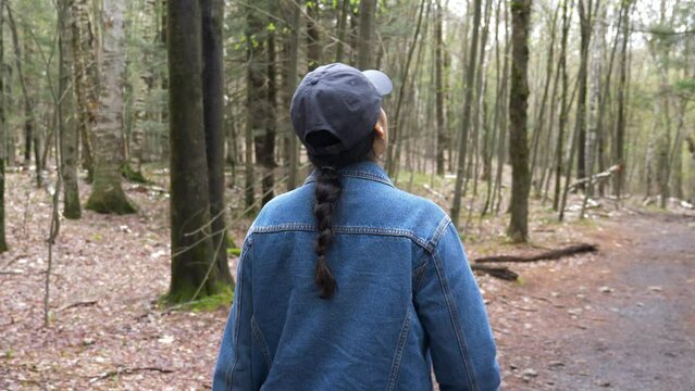 Young diverse woman wearing a long brunette braid jean jacket and grey baseball cap hat walking hiking through hike trail path in dense forest upstate new york during fall spring time 4k slow motion