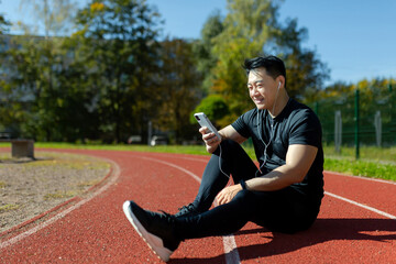Obraz premium Asian runner resting after physical active exercise, man with headphones and phone watching training video and listening music audio podcasts, sitting in stadium.