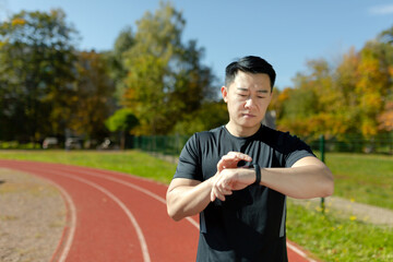 Asian runner checking heart rate and progress on fitness bracelet smartwatch, serious man looking thoughtful, running and doing fitness in stadium.