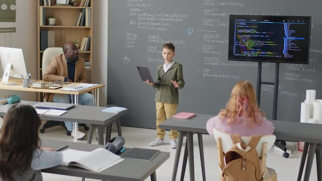Full shot of Caucasian elementary age boy standing in front of class in school holding laptop and giving presentation on programming at daytime