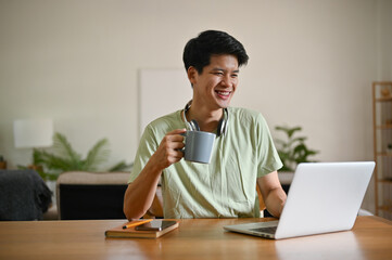 A happy young Asian man is sipping coffee while looking down at his laptop screen at a table
