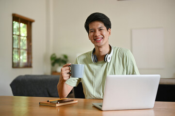 A happy Asian man sits at a table with a coffee cup in his hand and a laptop on a table.