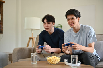 Two joyful young Asian male friends are sitting on a sofa and enjoying playing a video game