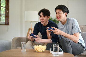 Fototapeta na wymiar Two excited Asian male friends are enjoying playing video games on a couch in the living room