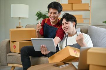 Fototapeta na wymiar A cheerful Asian gay couple checks their online product orders on a tablet, working at home together