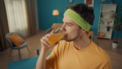 Profile portrait of a man enjoying drinking juice from a glass close up. The man in a tracksuit and yellow sports headband in a living room with sports equipment on the floor. - Powered by Adobe