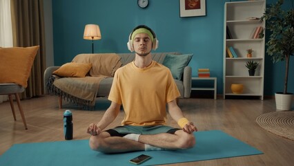 A young man sits cross legged in a lotus position with his eyes closed on a sports mat next to a smartphone. A man in a tracksuit and white wireless headphones is doing yoga at home.