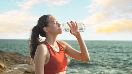 young woman drinks water from a bottle against the backdrop of the ocean