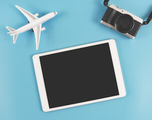  flat lay of digital tablet with blank black  screen, digital camera and  airplane model  isolated on blue background. travel planning concept.