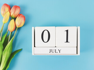 flat lay of wooden calendar with date July 01 on blue  background with orange and yellow tulips,...
