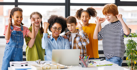 Teacher and cheerful  schoolkids   looking at laptop screen and celebrate   successful completion of collective school work   during online lesson