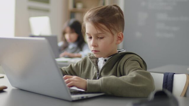 Medium shot of elementary age boy sitting at desk in school classroom working on project in laptop at daytime