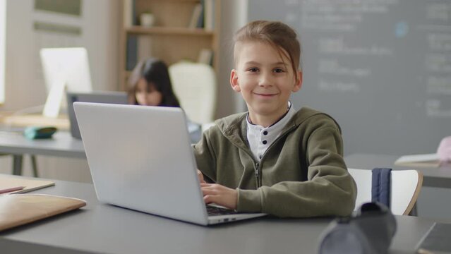 Slowmo portrait of Caucasian elementary age boy sitting at desk in school classroom in front of laptop looking at camera and smiling