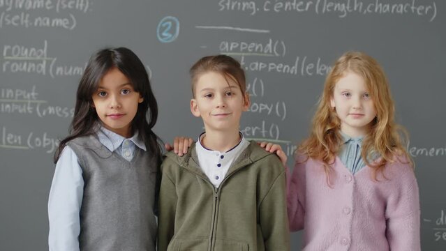 Slowmo portrait of three ethnically diverse elementary age students standing in front of blackboard looking at camera and smiling during programming lessonSlowmo portrait of three ethnically diverse e