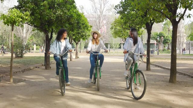 Young group of multiracial women having fun riding bike together in the park. Three smiling female friends enjoying time together on summer vacation. Friendship and holidays concept.