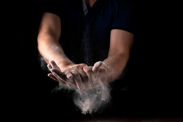 Fototapeta na wymiar A cloud of flour scatters from male hands on a black background. Cooking concept.