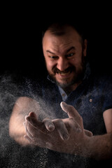 Obraz na płótnie Canvas The man claps his hands emotionally with anguish. A cloud of flour scatters from male hands on a black background. Cooking concept.