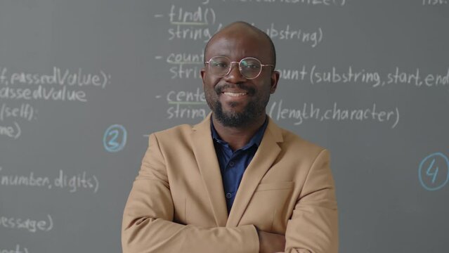 Medium close up portrait of male African American teacher standing in front of blackboard looking at camera and smiling in programming classroom at daytime