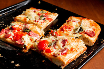 Sliced square pieces of pizza, pie, with tomatoes, sausage, cheese cooked in the oven on a black baking sheet.