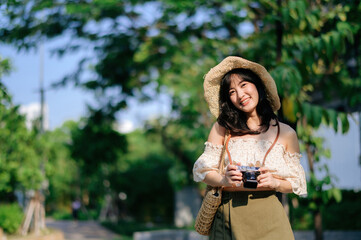 Portrait of asian young woman traveler with weaving hat and basket and a camera on green public park nature background. Journey trip lifestyle, world travel explorer or Asia summer tourism concept.