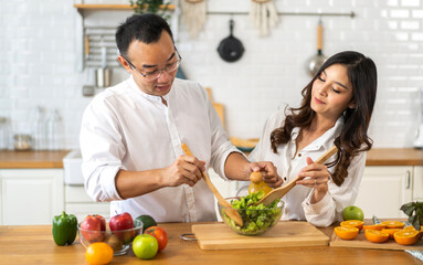 Young asian family couple having fun cooking and preparing cook vegan food healthy eat with fresh vegetable salad on counter in kitchen at home.Happy couple looking to preparing food
