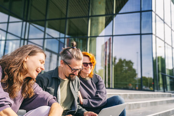 Group of happy smiling young adult people sitting near office building looking at laptop screen...