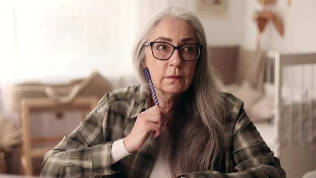 Portrait of beautiful gray haired senior woman thinks questioningly about difficult choice choose imagine plan in mind has doubts create idea at desk at home workplace