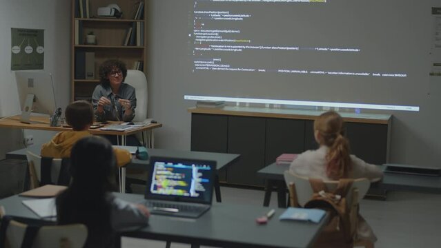 Full shot of Caucasian female teacher sitting at desk in classroom having discussion with elementary age students during programming lesson