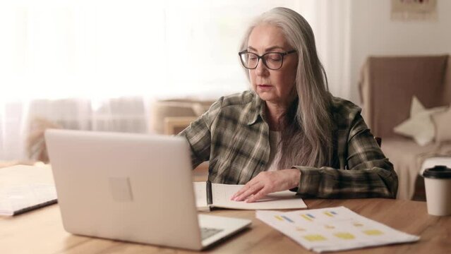 Focused gray haired senior woman studying online at home Curious mature female make notes in notebook while looking at laptop computer screen Distance remote education for person on pension