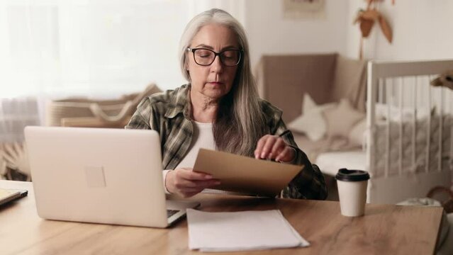 Portrait of beautiful gray haired senior woman checking mail correspondence open envelope and reading document sitting next to laptop computer at home workplace