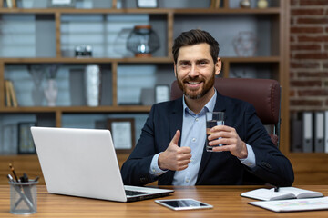 Portrait of successful mature businessman inside office at workplace, senior experienced man smiling and looking at camera, using computer at work, drinking water and showing thumbs up encouraging.