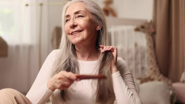 Portrait of beautiful senior woman looking at mirror and combing her long gray hair with wooden brush in the morning at home Concept of self care and daily routine