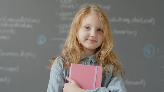 Slowmo portrait of Caucasian elementary school girl posing in front of blackboard in classroom holding notebook during programming lesson at daytime