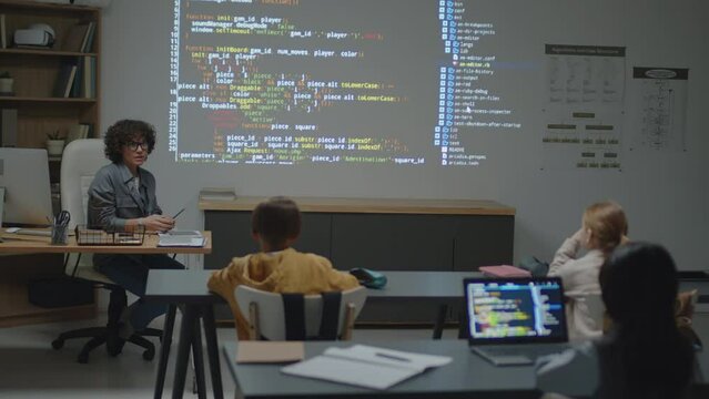 Full shot of Caucasian female teacher sitting at desk in classroom pointing at screen projection with code explaining it to elementary age students
