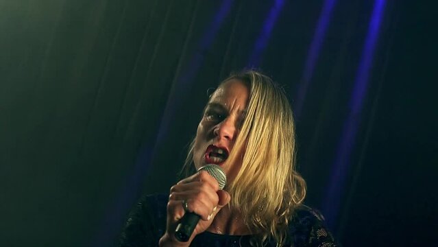 Handheld medium shot of a blonde talented singer during a concert, singing with a lot of emotion in her facial expressions and gestures with her microphone during a coloured strobe light