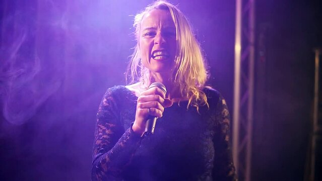 Handheld medium shot of a talented blonde singer performing and singing a song emotionally during a concert with a coloured light show in changing colours