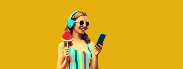 Summer portrait of happy smiling young woman model in headphones listening to music on smartphone with juicy lollipop or ice cream shaped slice of watermelon on yellow background