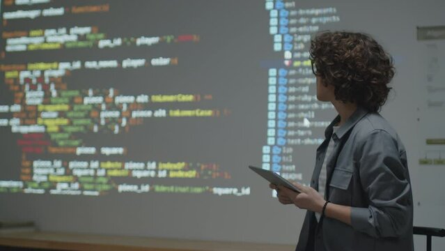 Medium shot of female Caucasian woman standing in front of class holding tablet in hands and giving lecture on programming showing example on screen projection