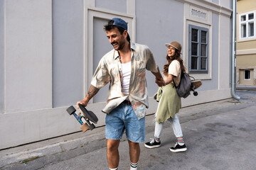 Fototapeta premium Tourists couple skateboarders riding skates. Happy young travelers longboarding together on sunset on pavement. Stylish man and woman in trendy outfit skating outdoors in touristic destination streets