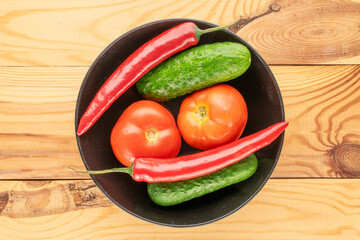 Two pods of hot red pepper, two tomatoes, two cucumbers with a black plate on a wooden table, macro, top view.