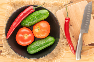 Two hot red pepper pods, two tomatoes, two cucumbers with a black tao oil with a knife and a wooden kitchen board, macro, top view.