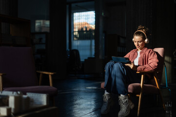 Young woman stting in armchair, listening music and working on digital tablet.