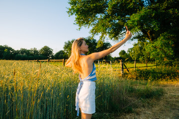 Naklejka na ściany i meble Young attractive cheerful woman making selfie photo using mobile phone at the countryside field of wheat in summer day at sunset.