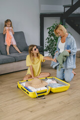 Mother with daughters packing yellow suitcase for travel. Family collecting clothes and accessories for trip in luggage. Happy and bored child getting ready for vacation.