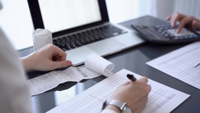 Two accountants use a calculator and laptop computer for counting taxes or revenue balance while rolls of receipts are waiting to be calculated. Business audit, and taxes concepts