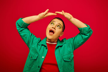 fashion and style, amazed and short haired woman in green outfit posing with raised hands on red background, looking up generation z, youth culture, vibrant backdrop, individuality, personal style