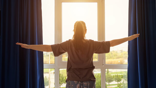 Brown-haired woman wearing t-shirt starts morning routine from simple exercises. Lady doing exercises looks at cityscape outside window, sunlight