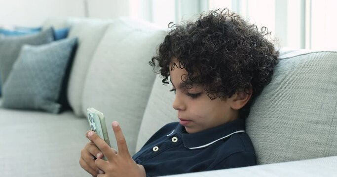 Cute curly-haired Italian boy rest on sofa with cellphone spend time play online game. Generation Alpha and modern wireless technology overuse. Parental control application for kids safety on internet