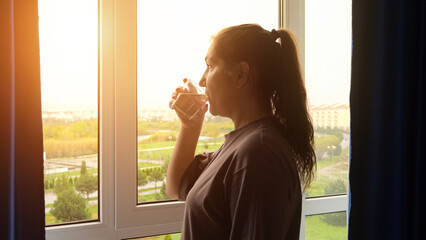 House-woman drinking water from glass looks at city outside window. Adult woman takes care of...