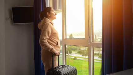 Brown-haired woman comes to window with black travel suitcase in apartment. Adult housewoman missing hometown prepares for long journey, sunlight
